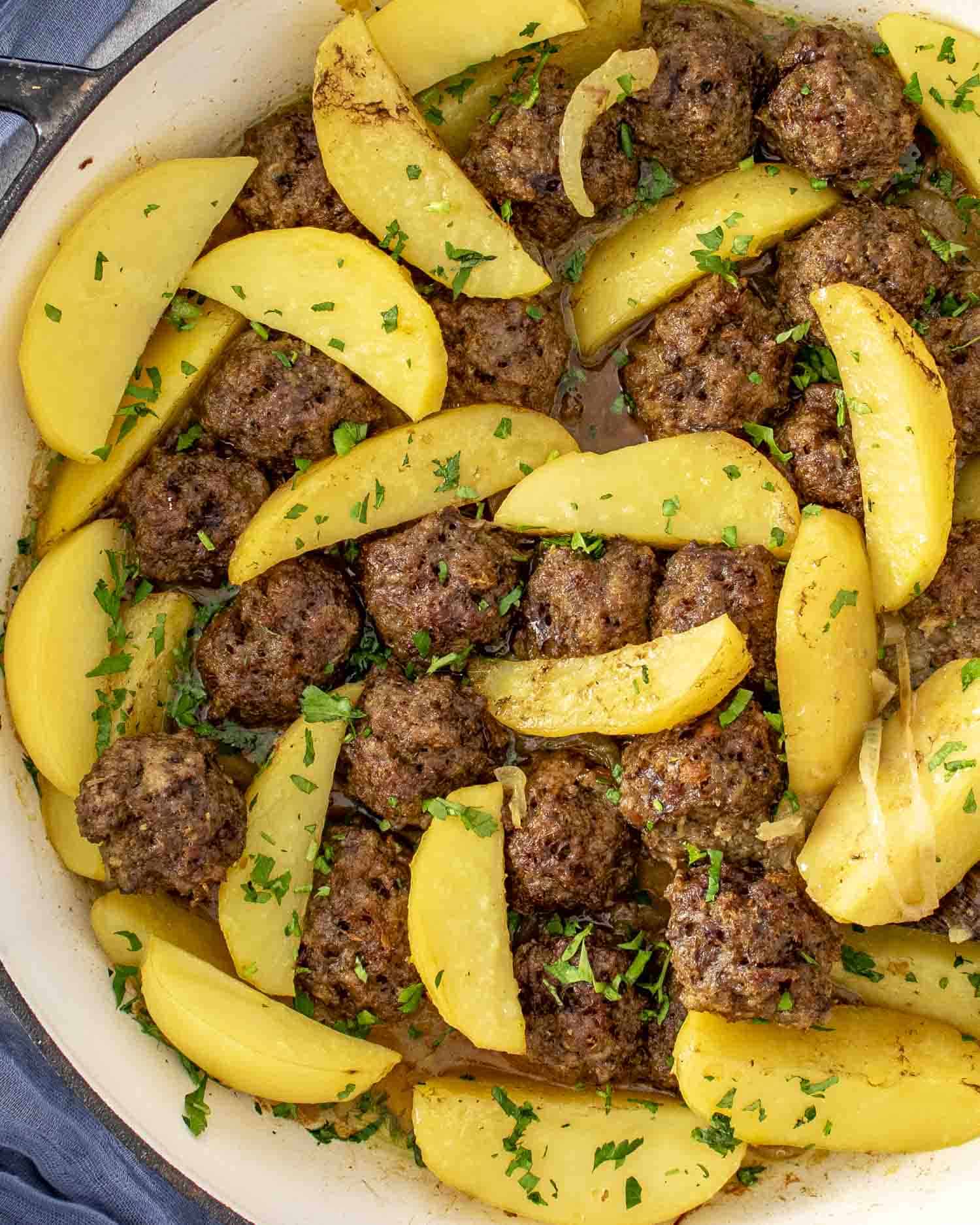A close-up of a large white pot filled with golden potato wedges and browned meatballs, garnished with fresh chopped parsley. The dish is saucy, vibrant, and comforting, showcasing a hearty one-pot meal.