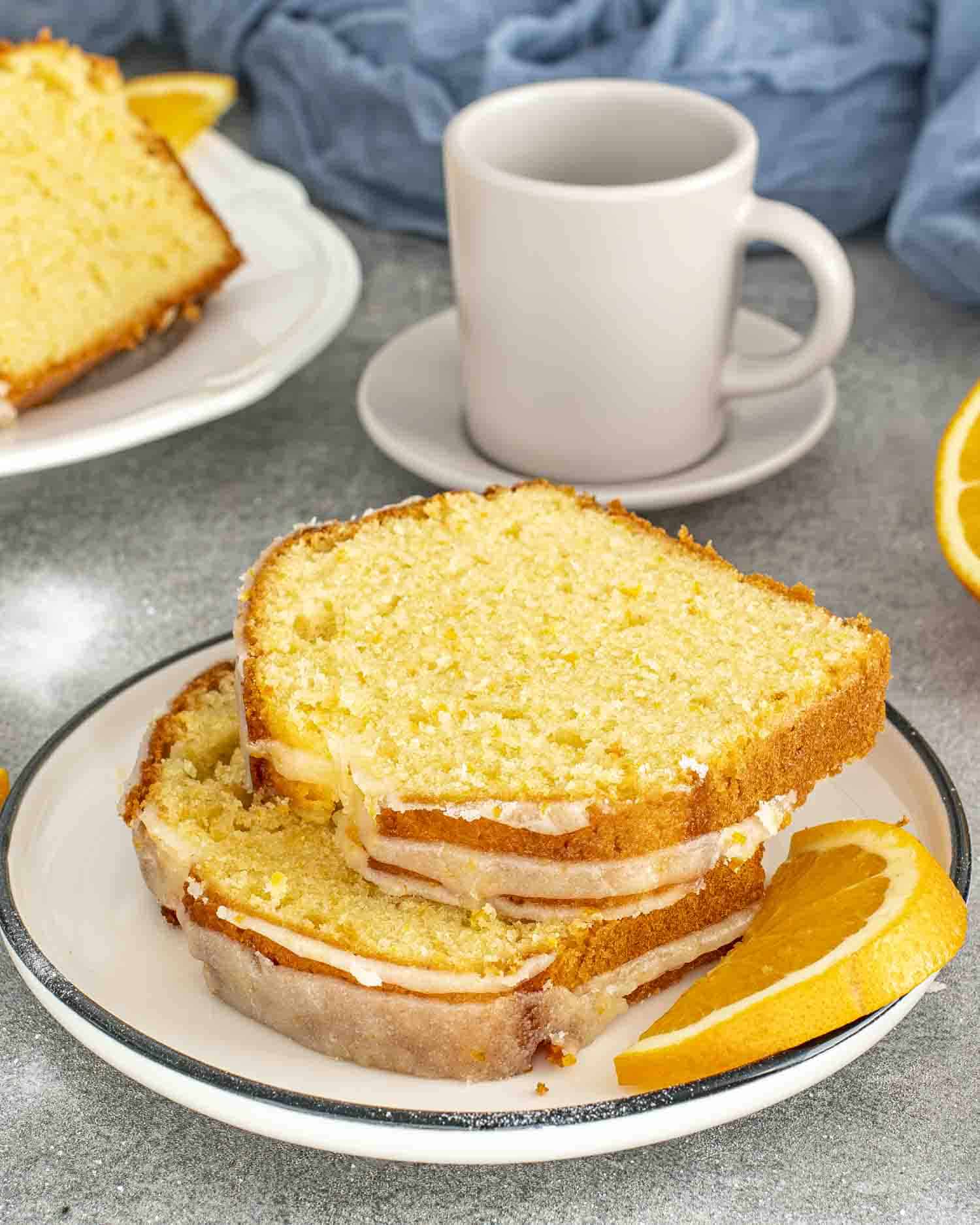 2 slices of orange cake drizzled with an orange glaze on a white plate with a cup of coffee in the background.
