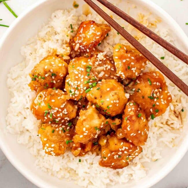 orange chicken on a bed of rice garnished with chives in a white bowl.