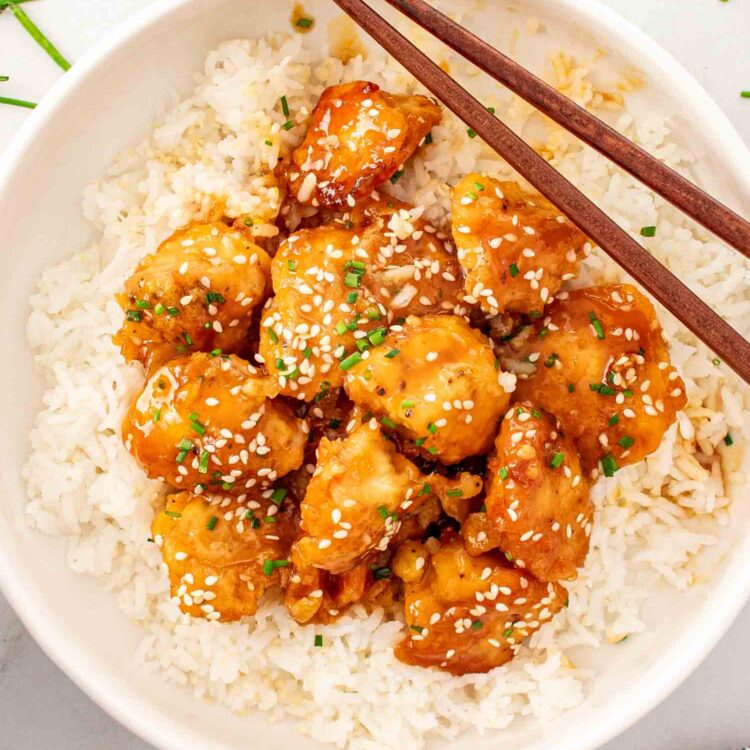 orange chicken on a bed of rice garnished with chives in a white bowl.