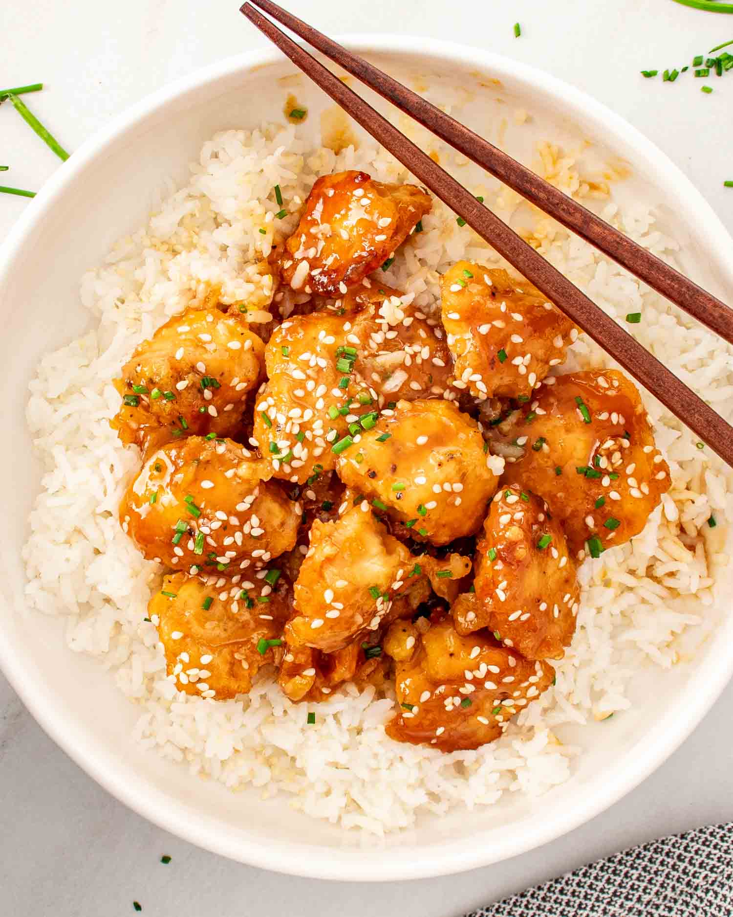 orange chicken on a bed of rice garnished with chives in a white bowl.
