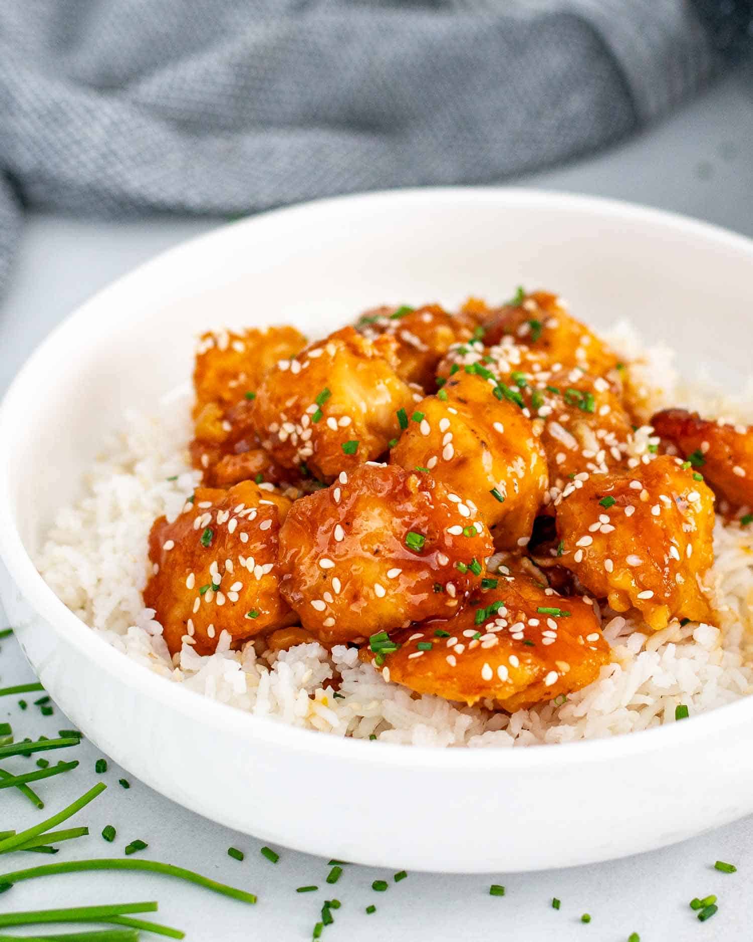 orange chicken on a bed of rice garnished with chives in a white bowl.