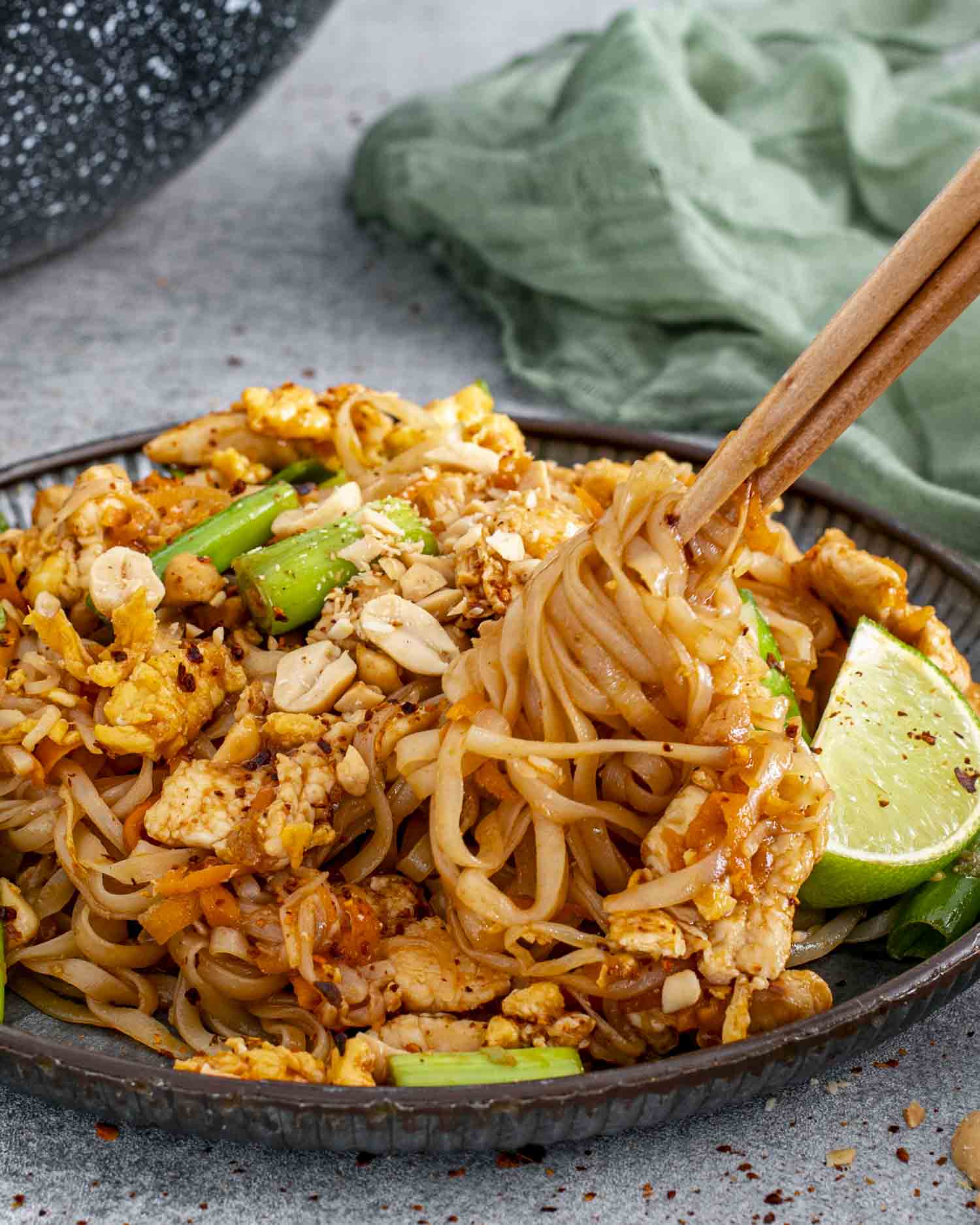 A plate of Pad Thai with rice noodles, tender chicken, scrambled eggs, green onions, and crushed peanuts. Chopsticks are lifting a portion of noodles, while a lime wedge adds a pop of color on the side.