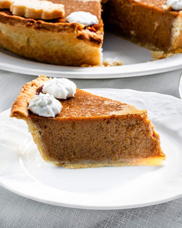 a slice of pumpkin pie on a white plate with 2 dollops of whipped cream