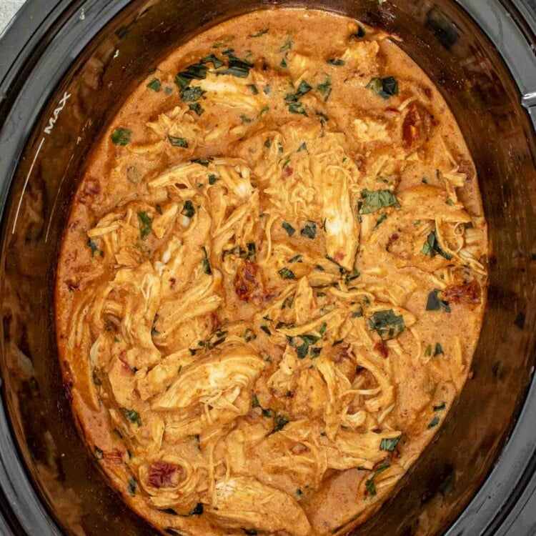Close-up of shredded chicken in a creamy tomato Parmesan sauce inside a slow cooker, topped with chopped basil.