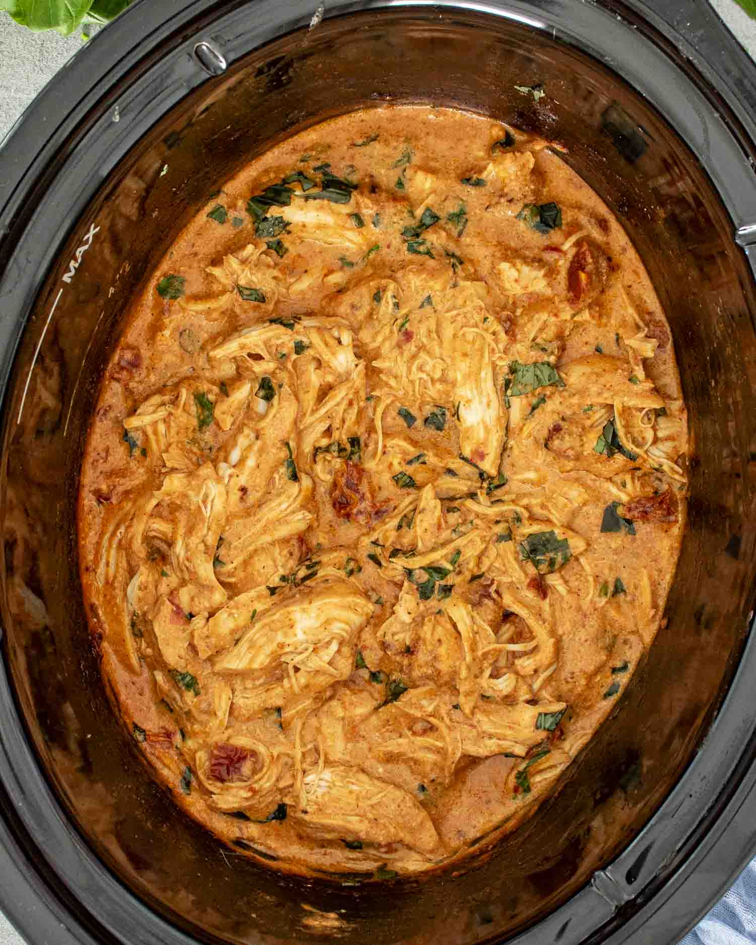 Close-up of shredded chicken in a creamy tomato Parmesan sauce inside a slow cooker, topped with chopped basil.