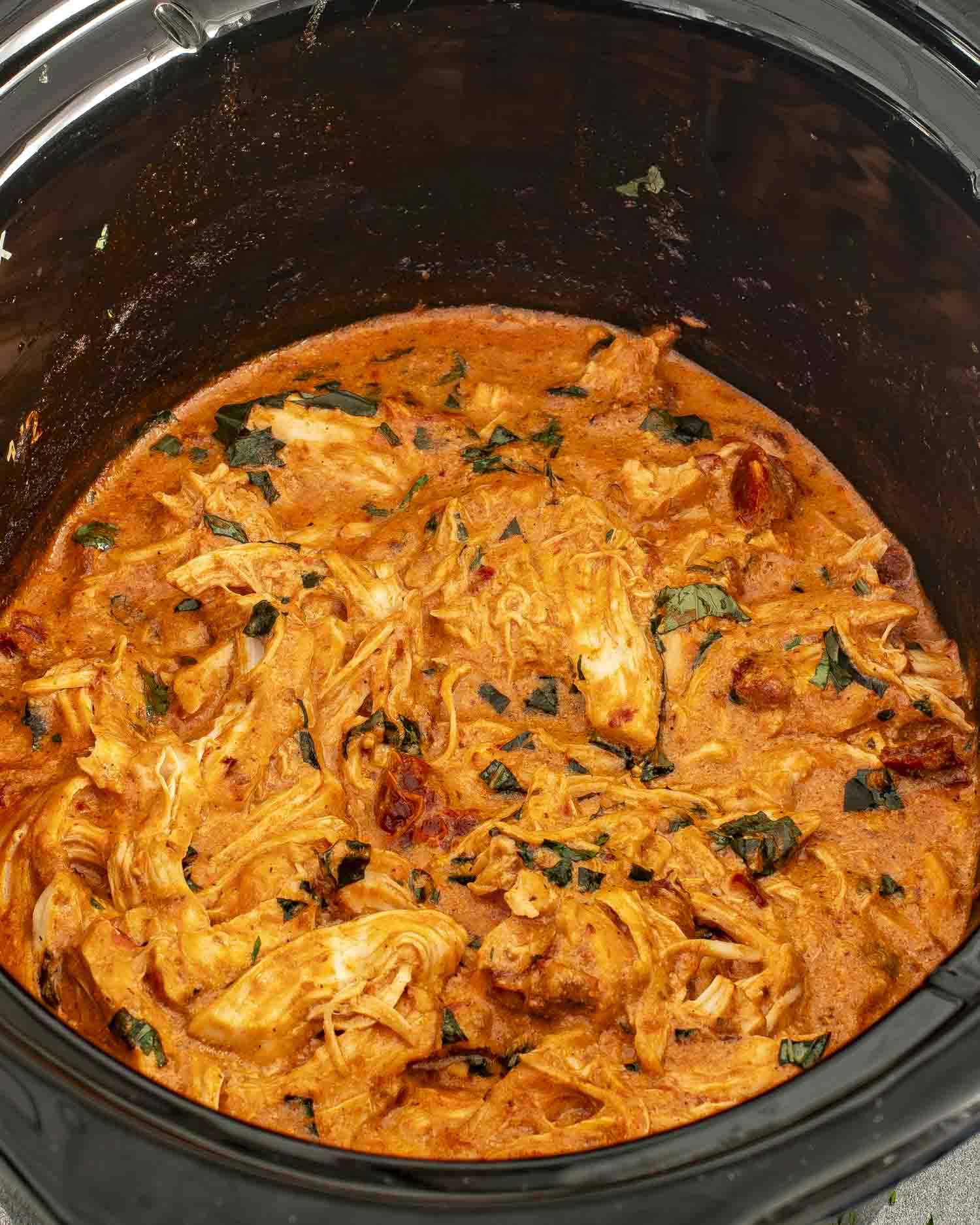 Close-up of shredded chicken in a creamy tomato Parmesan sauce inside a slow cooker, topped with chopped basil.