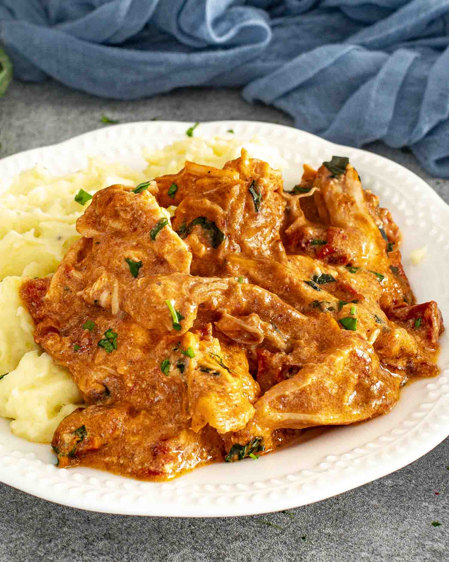 Creamy slow cooker Marry Me Chicken on mashed potatoes, topped with fresh basil on a white scalloped plate.