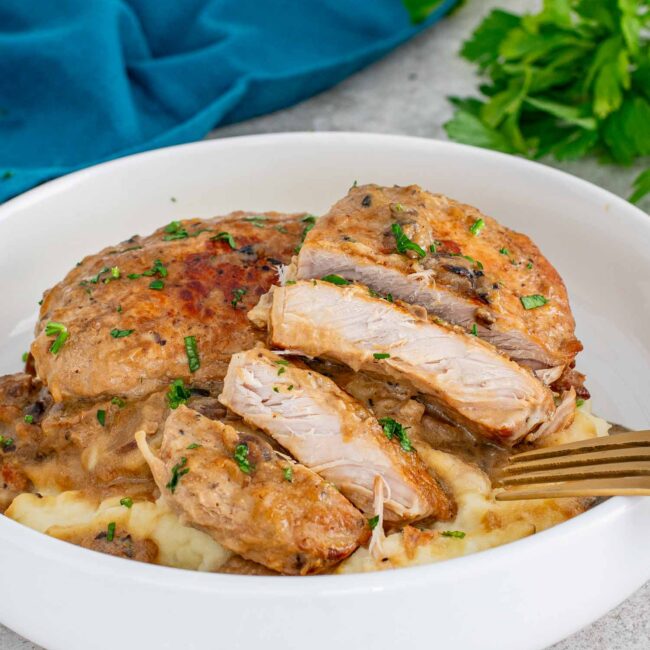 2 pork chops with mushroom gravy made in the slow cooker on a bed of mashed potatoes on a white plate.