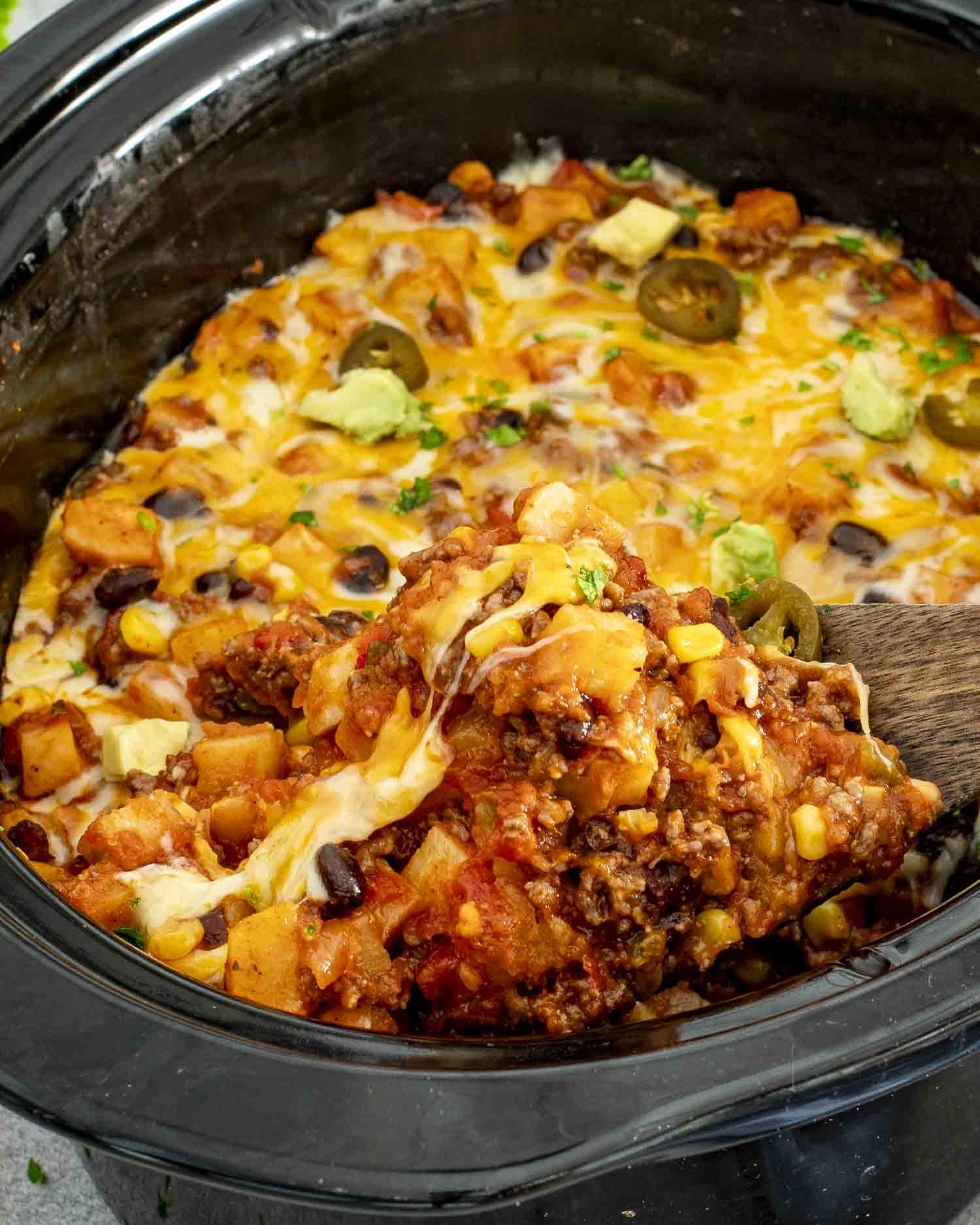 Slow cooker taco casserole with melted cheese, potatoes, corn, beans, and beef being scooped with a wooden spoon.