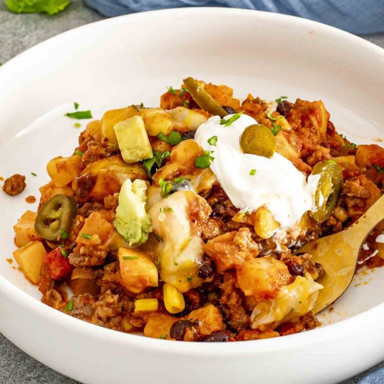 Close-up of taco casserole served in a white bowl, topped with sour cream, avocado, jalapeños, and melted cheese.
