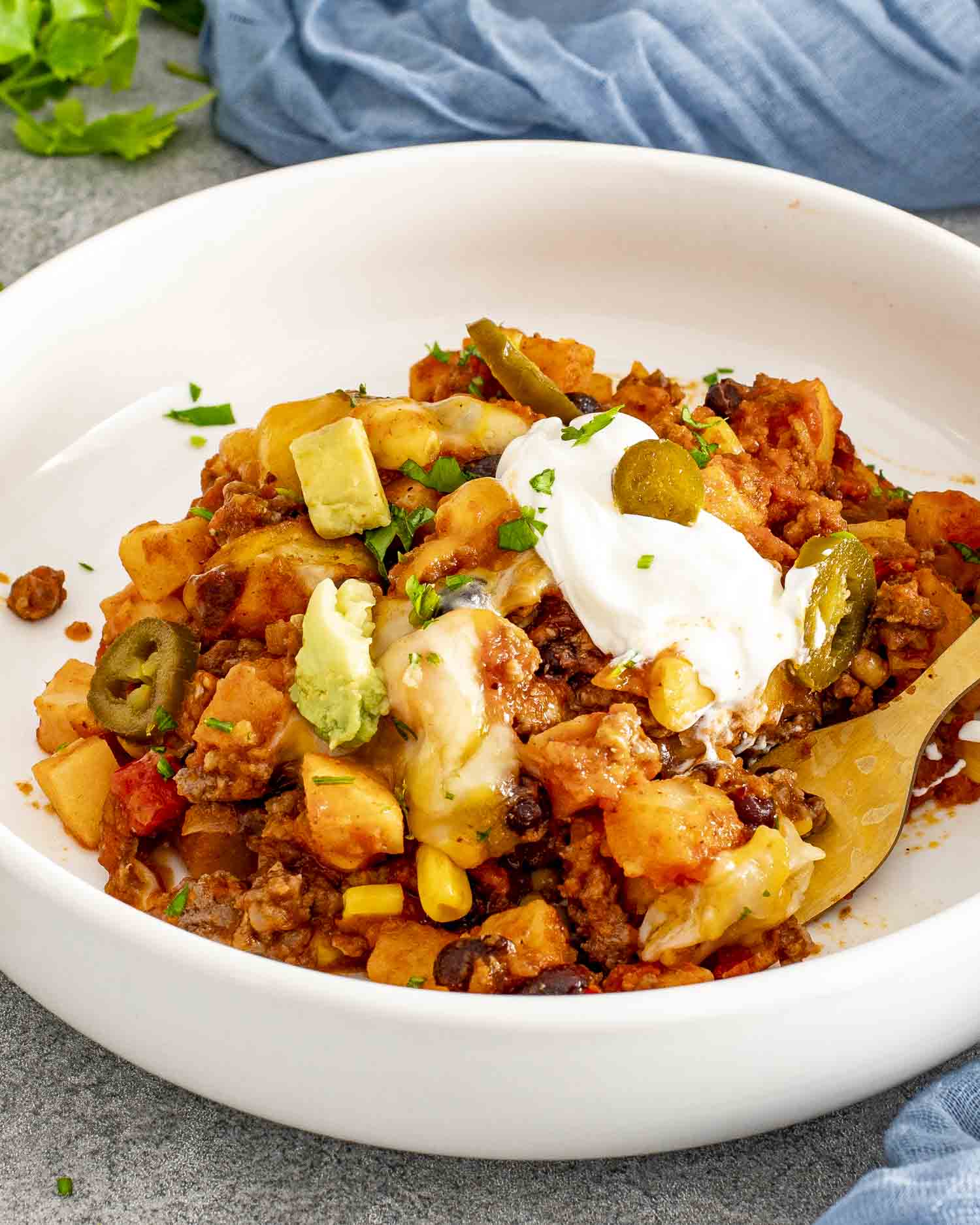 Close-up of taco casserole served in a white bowl, topped with sour cream, avocado, jalapeños, and melted cheese.