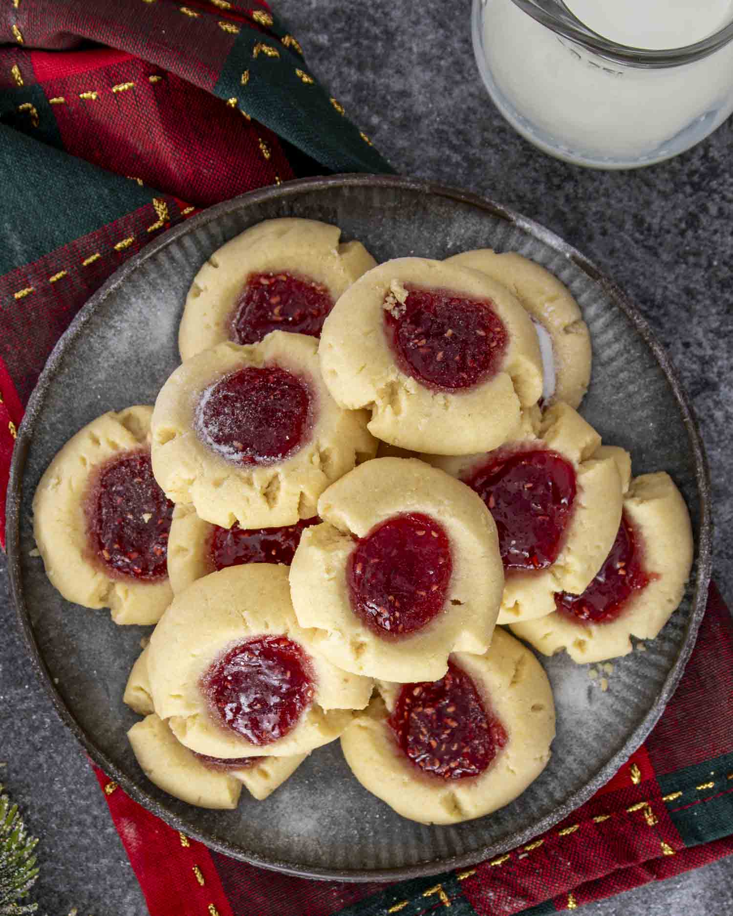 A full plate of buttery thumbprint cookies with jam centers, served on a dark surface with holiday napkin and milk.
