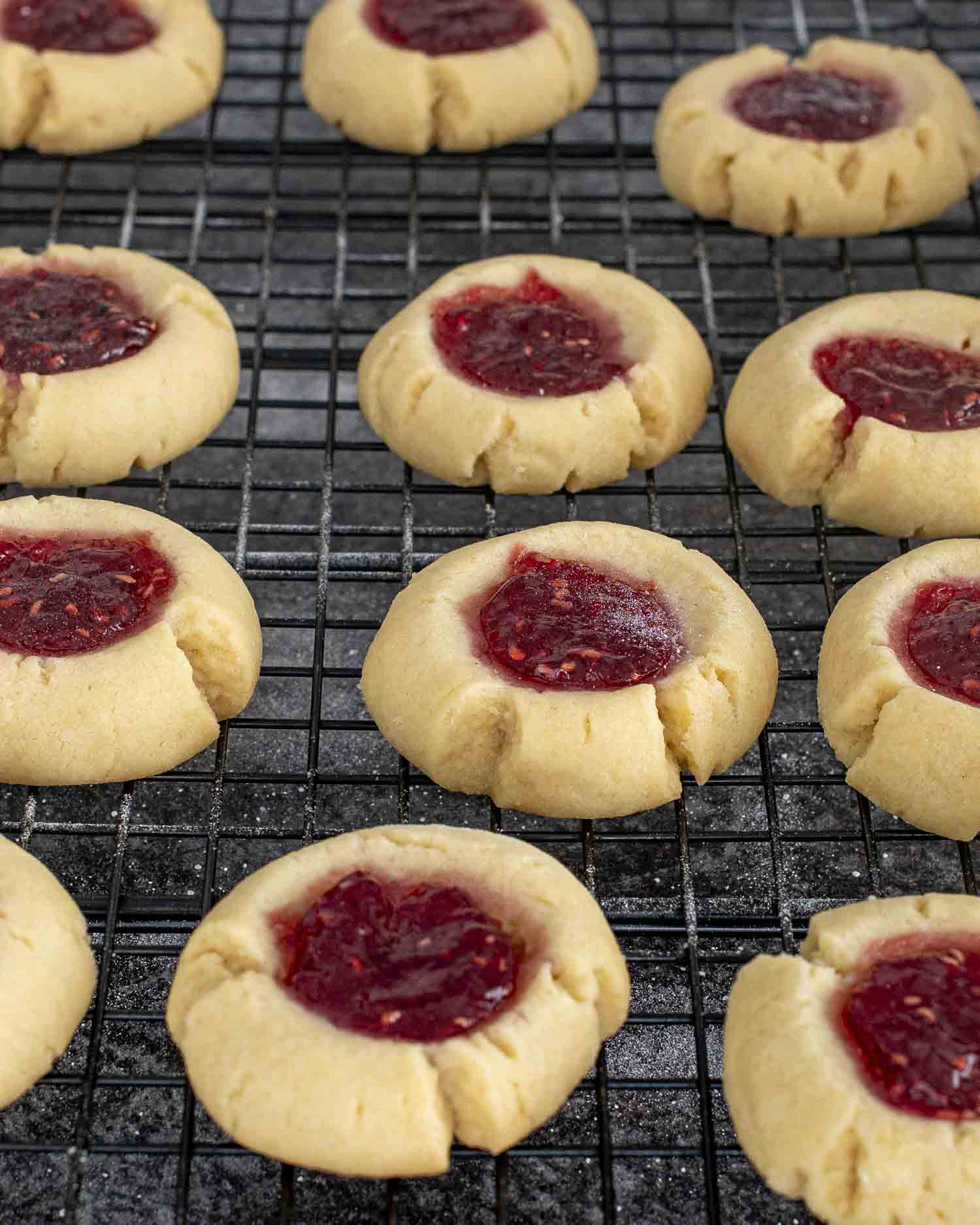 Freshly baked thumbprint cookies with raspberry jam centers cooling on a wire rack, golden edges and glossy fruit filling.