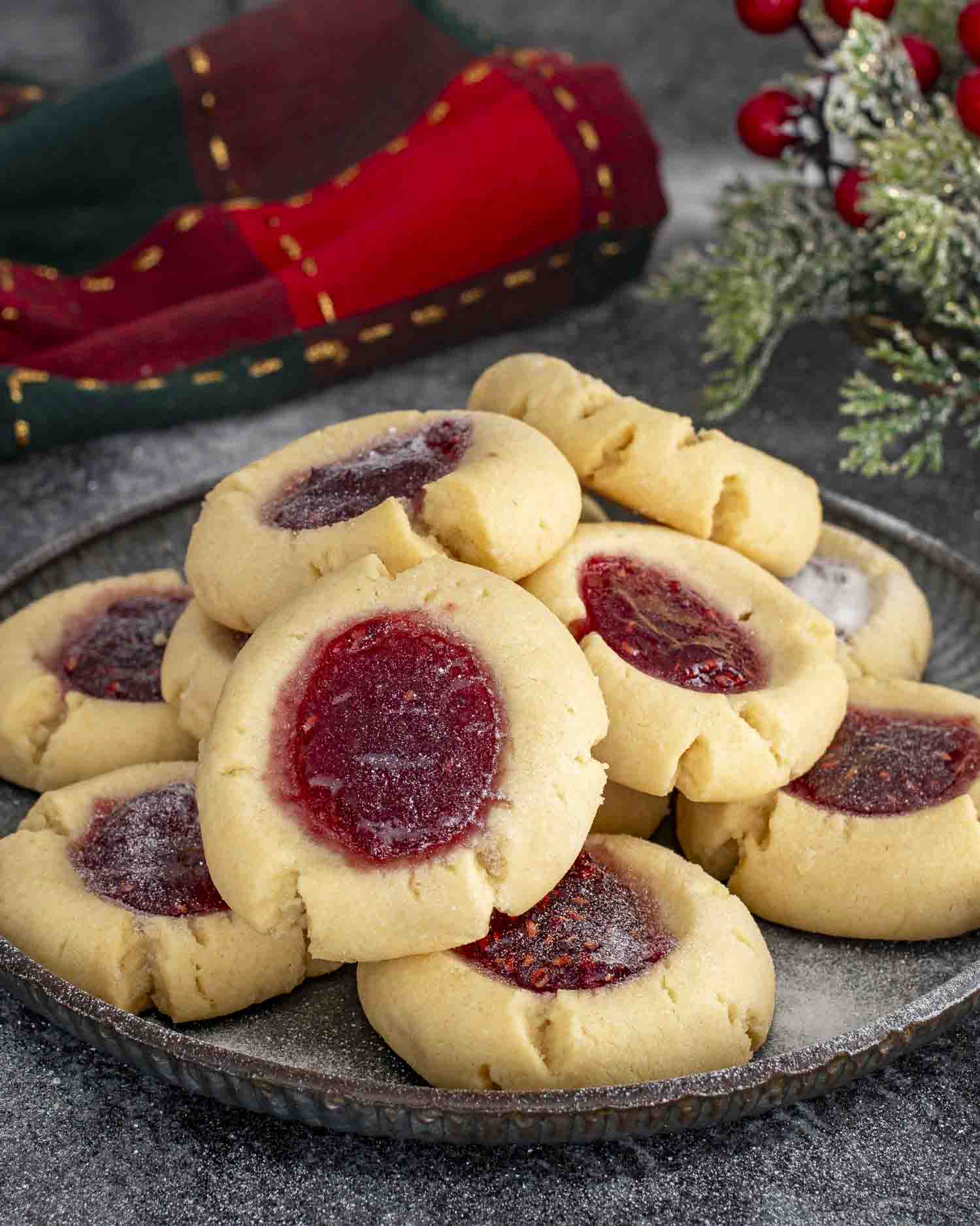 A plate stacked with buttery thumbprint cookies filled with red jam, lightly dusted with sugar, ready for holiday sharing.