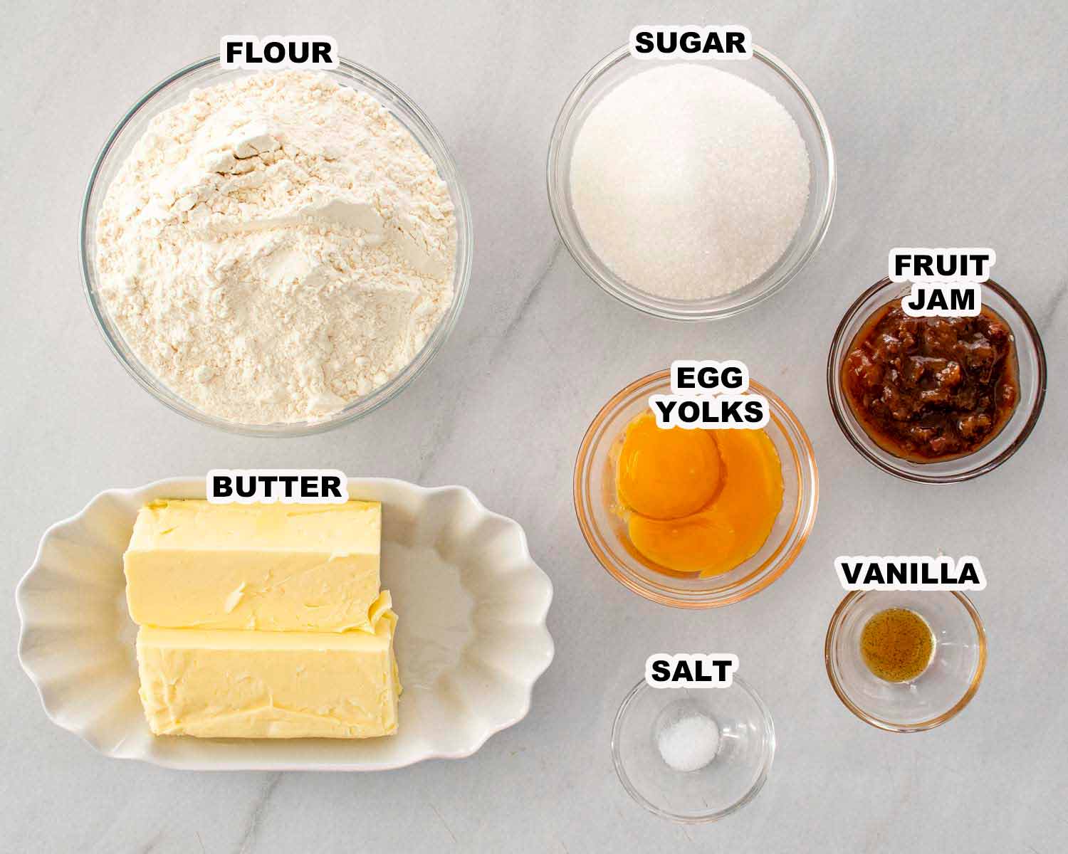 Glass bowls filled with butter, flour, sugar, egg yolks, vanilla, salt, and jam, all measured and labeled for thumbprint cookies.