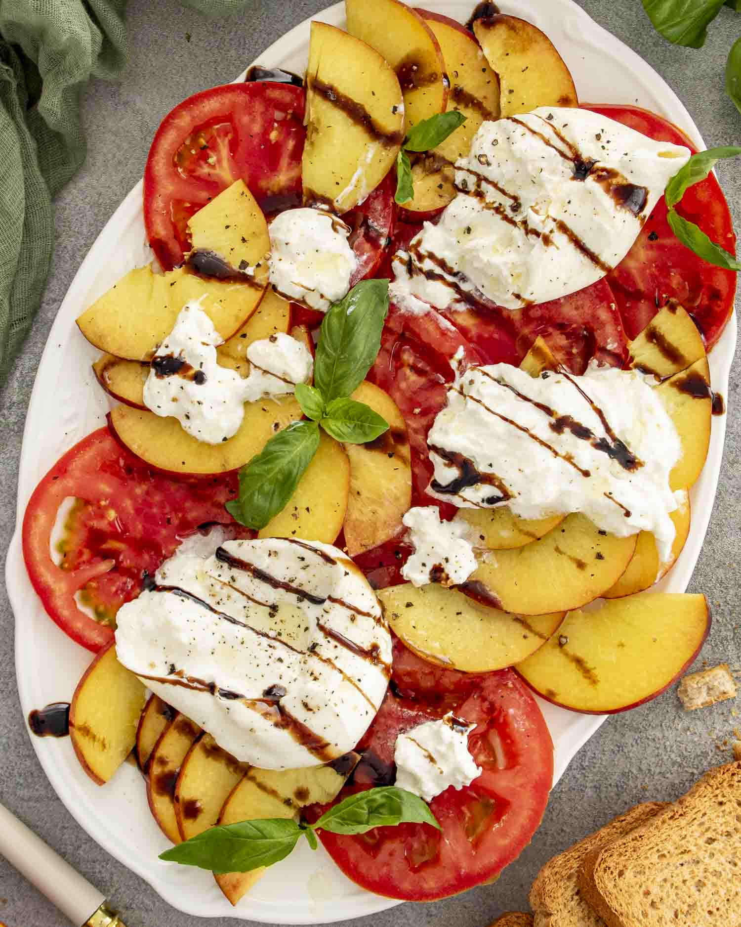 Colorful salad featuring ripe tomatoes, juicy peach slices, torn burrata cheese, basil leaves, and balsamic glaze on a large white platter.