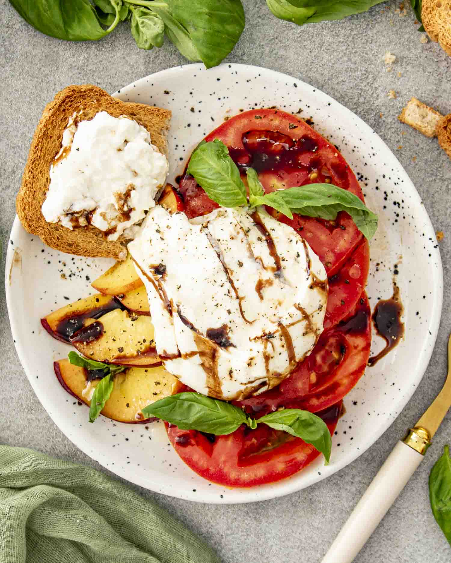 Overhead view of sliced tomatoes and peaches with burrata and balsamic glaze, served beside toasted bread on a speckled white plate.