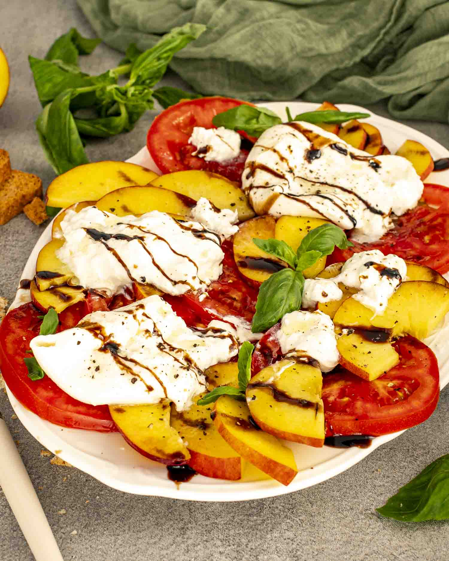 A platter filled with vibrant red tomatoes, golden peaches, torn burrata, fresh basil leaves, and rich balsamic glaze drizzled over everything.