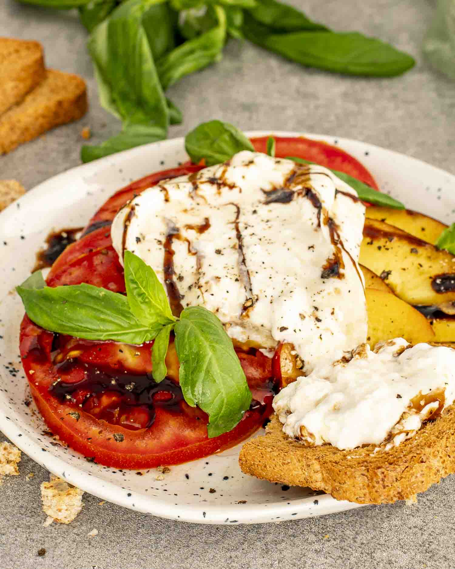Sliced tomatoes and peaches layered on a plate, topped with creamy burrata, balsamic drizzle, fresh basil, and crisp toasted bread.