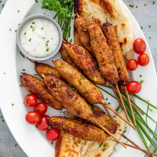 chicken kebabs drizzled with tzatziki sauce on a plate alongside some pita bread and cucumbers.