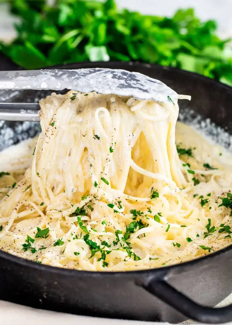 a pair of tongs lifting pasta alfredo from a cast iron pan