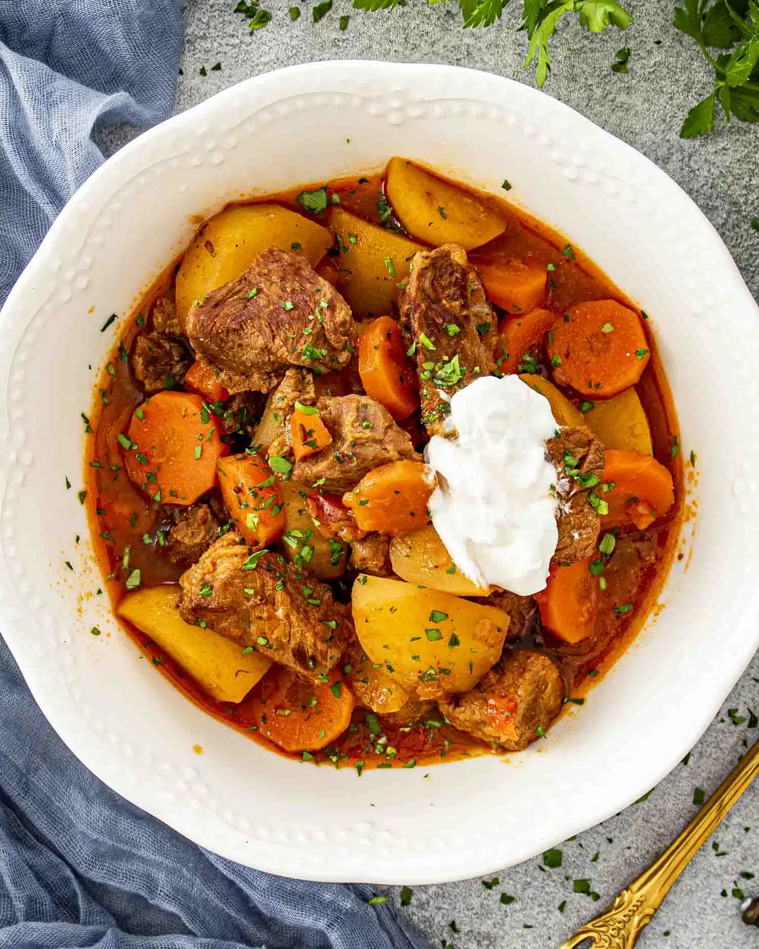 A top-down shot of a bowl of Beef Goulash served in a white, textured ceramic dish. Tender chunks of beef, golden potatoes, and sliced carrots are coated in a rich, paprika-infused broth. A dollop of sour cream sits on top, melting slightly into the hearty stew, while freshly chopped parsley adds a pop of color.