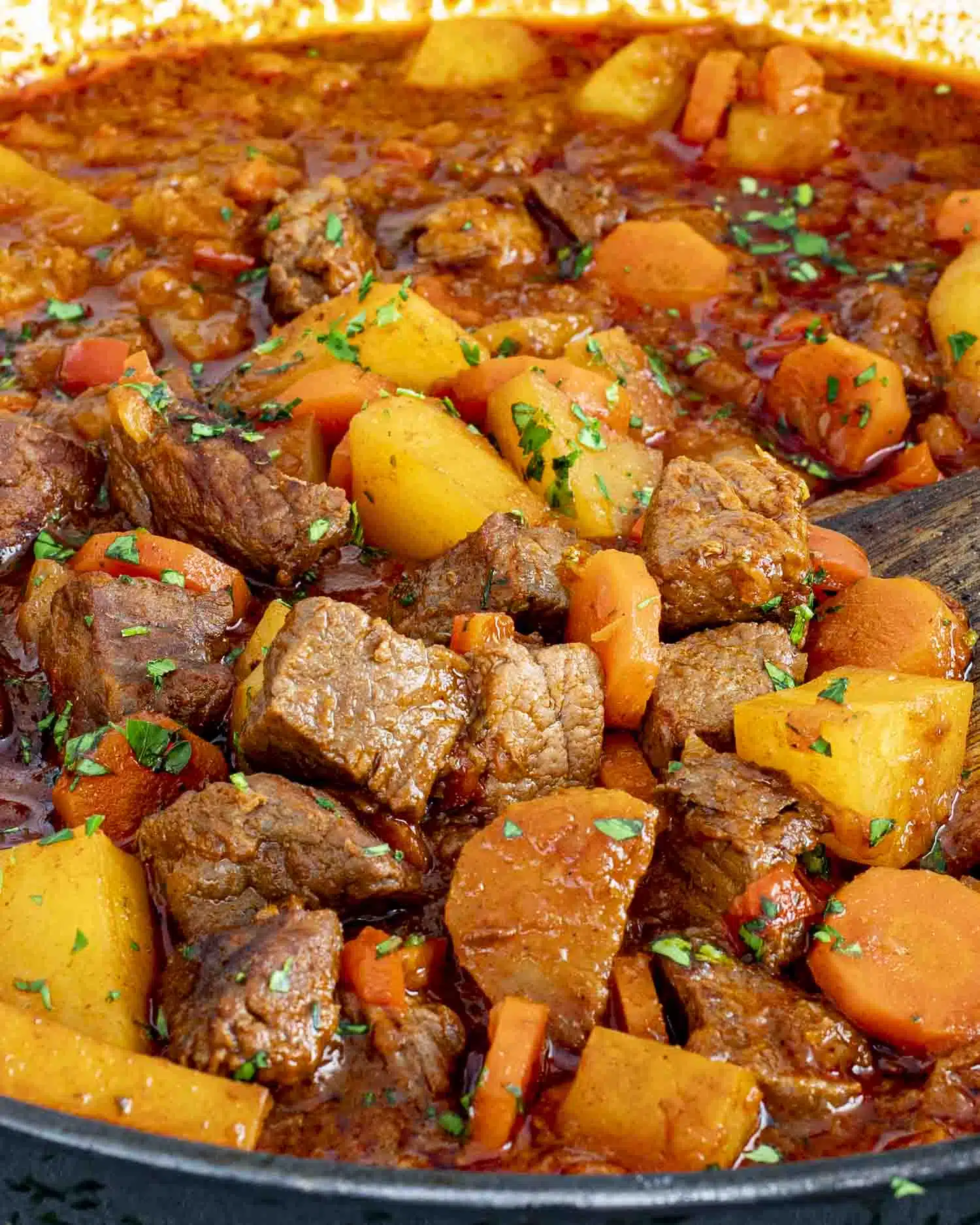 A close-up shot of a hearty Beef Goulash in a large Dutch oven. Tender chunks of beef, golden potatoes, and sliced carrots are simmered in a rich, deep-red paprika-infused broth. The dish is garnished with freshly chopped parsley, adding a pop of green. 