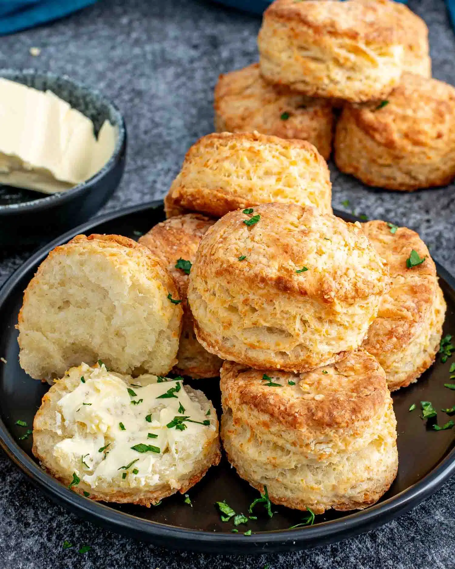a few buttermilk parmesan biscuits on a black plate with one cut in half and spread with butter.