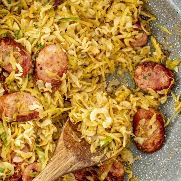 a big skillet with freshly made cabbage and noodles with sausage with a wooden spoon inside.