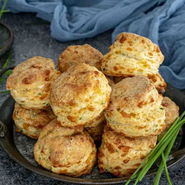 Freshly baked cheddar chive biscuits stacked high on a dark tray, showing golden flaky layers and melted cheddar throughout.