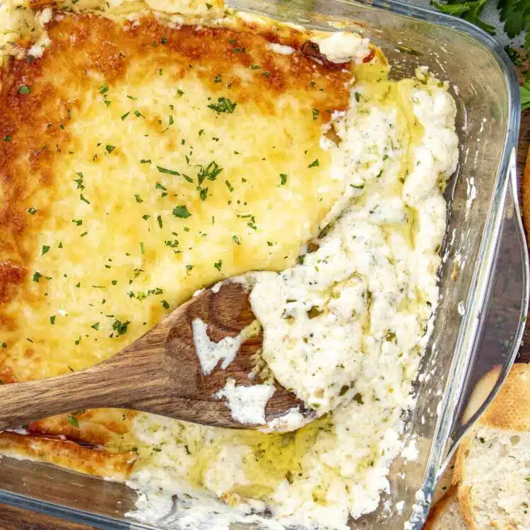 Creamy, cheesy roasted garlic dip with a golden top, partially scooped with a wooden spoon, served alongside crusty bread.