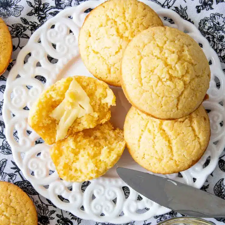 a cornbread muffin broken in half with butter on a plate with a few other cornbread muffins.