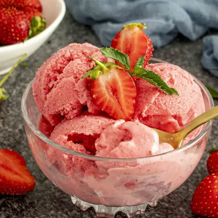 Scoops of frozen strawberry yogurt in a glass bowl topped with fresh strawberries and mint, golden spoon inside.