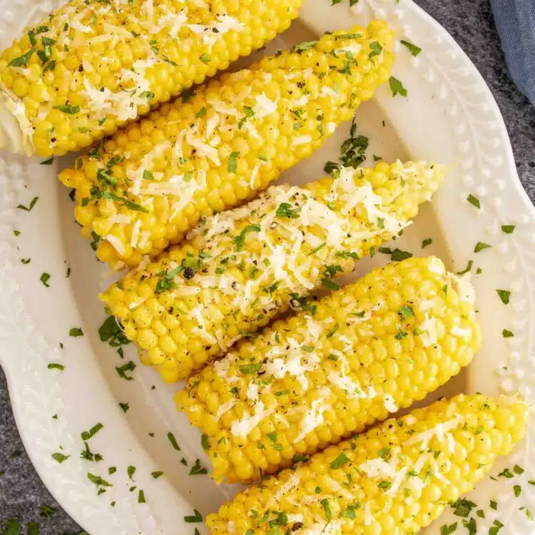 Overhead view of five corn cobs smothered in garlic butter, Parmesan, and parsley on a white serving dish.