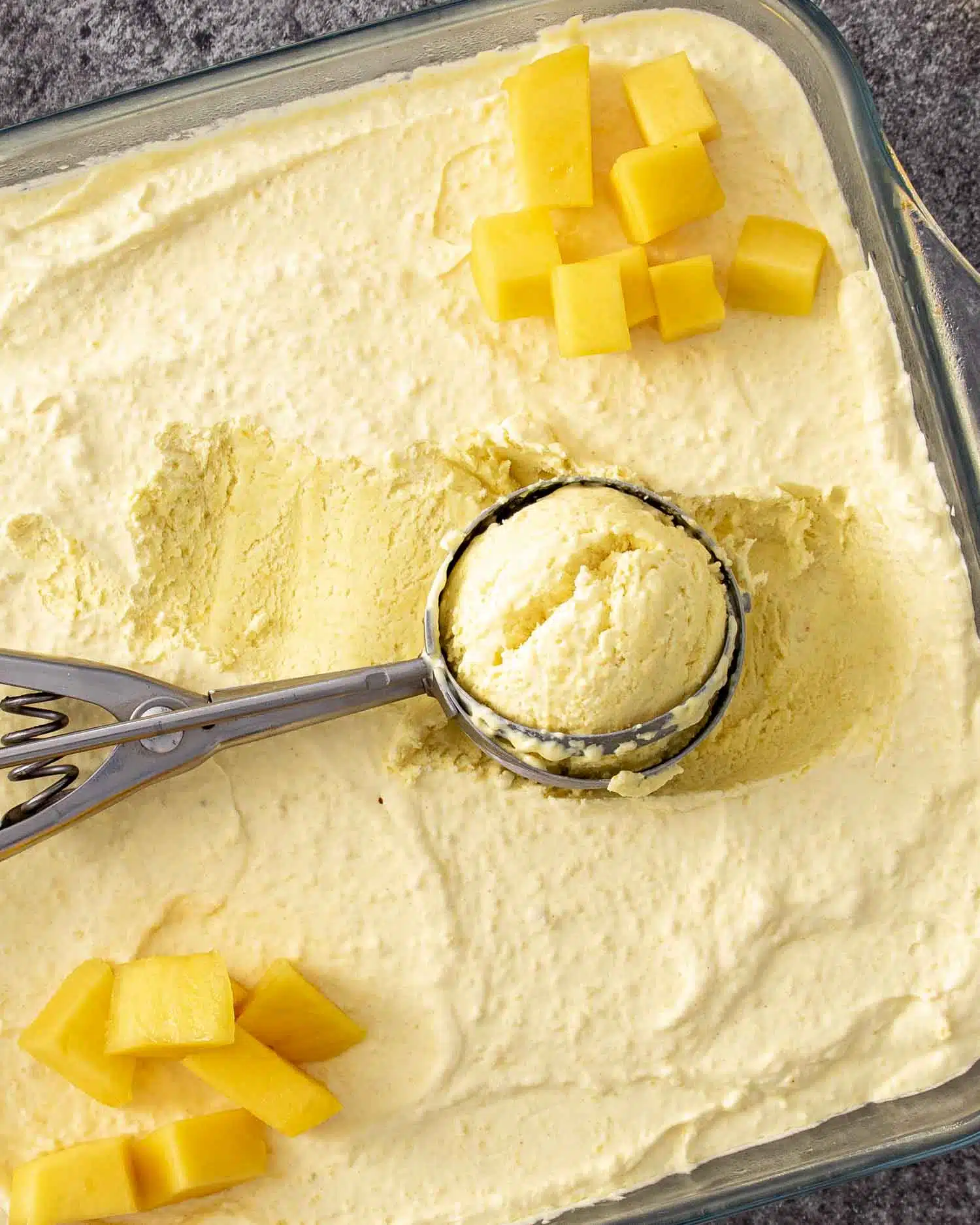 Overhead view of frozen mango ice cream being scooped, topped with bright mango chunks in a clear dish.