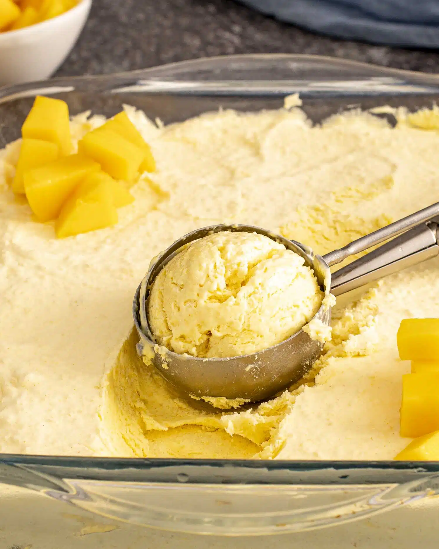 Glass container of no churn mango ice cream, with a scoop lifted and mango chunks scattered on top.