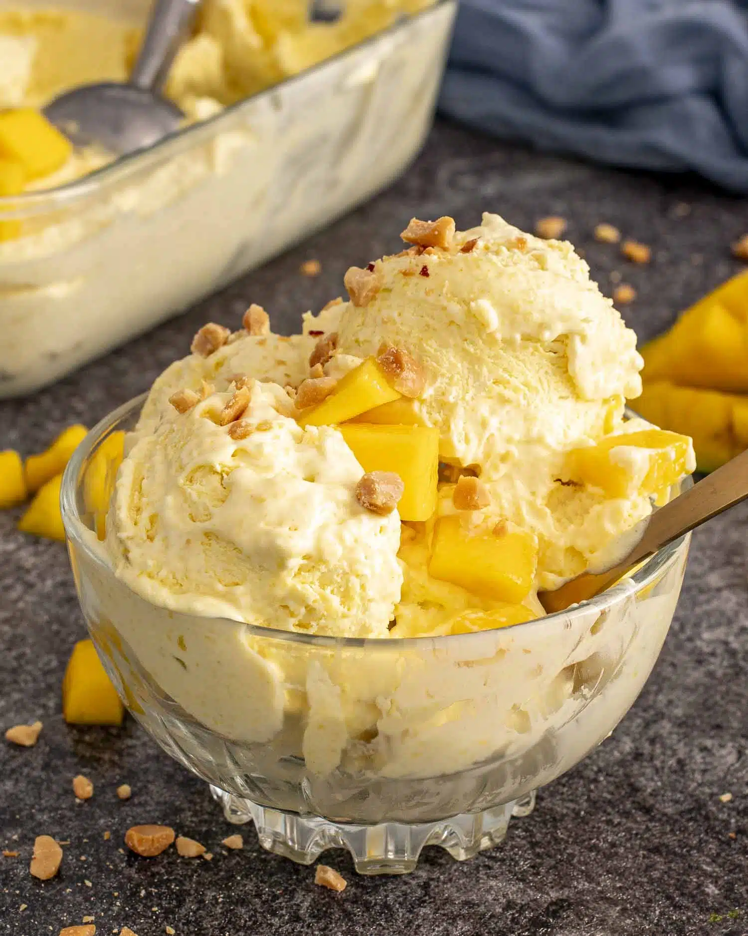 Scoops of creamy mango ice cream in a glass bowl, topped with chopped mango and crushed nuts, spoon inside.