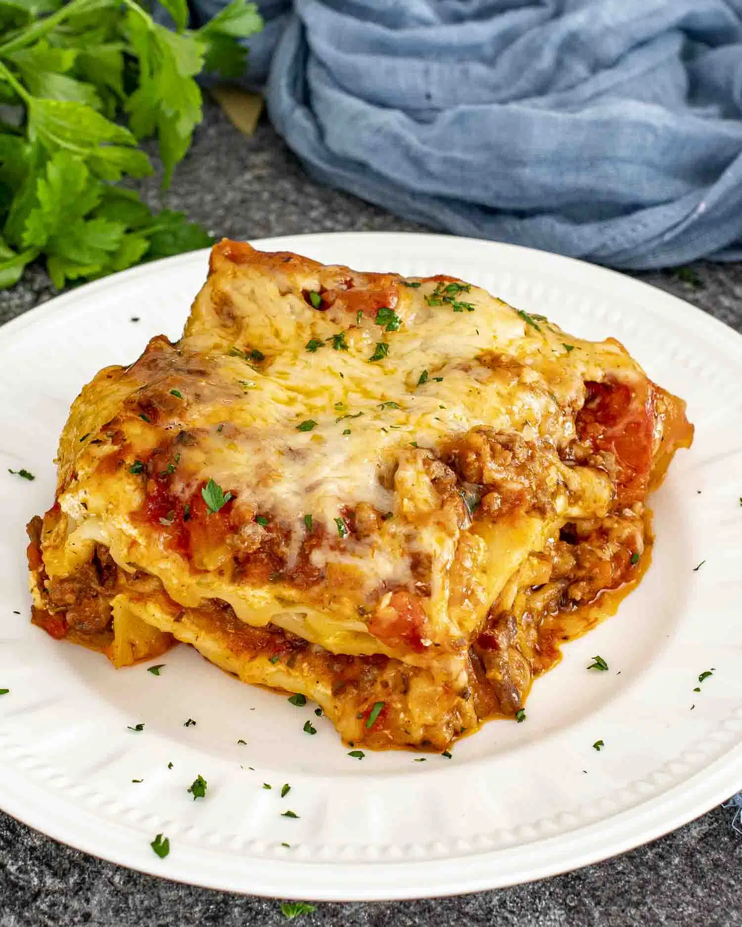 a slice of lasagna on a white plate garnished with a bit of parsley.