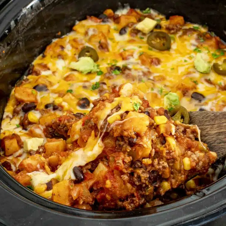 Slow cooker taco casserole with melted cheese, potatoes, corn, beans, and beef being scooped with a wooden spoon.
