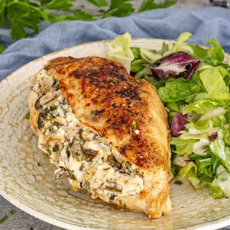 A stuffed chicken breast with a creamy spinach and artichoke filling, served with a fresh green salad on a speckled plate.