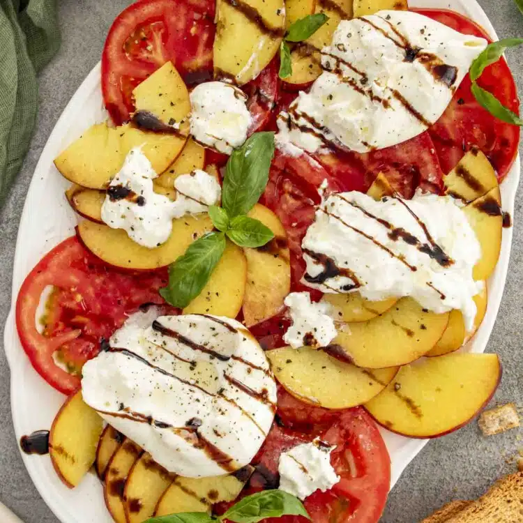 Colorful salad featuring ripe tomatoes, juicy peach slices, torn burrata cheese, basil leaves, and balsamic glaze on a large white platter.
